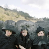 Four children dressed in dark clothing and hats stand closely together in front of a stone wall outdoors. The scene has a calm, posed feeling with a muted natural background.