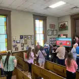 People gather and talk inside a historic building. A presentation screen and colorful windows are visible in the background. 