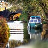 Hausboot auf einem Kanal umgeben von Bäumen, mit einer Brücke aus Stein im Hintergrund.