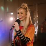 Una mujer sonriente con cabello largo y rubio que sostiene un micrófono como si estuviera a punto de comenzar a cantar.