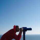Side on view of a man in a red coat bending down to look through a telescope pointed out of right of frame.