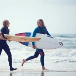 Zwei Personen in Neoprenanzügen, die Surfboards halten, laufen durch das Wasser über den Sand an einem Strand.