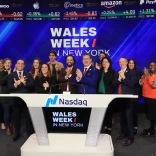 A group of people stand on the Nasdaq stage clapping and smiling during a Wales Week in New York event, with stock tickers displayed above them.