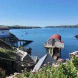 A small coastal harbour with calm blue water, a red topped lifeboat moored by a slipway, white buildings on the shoreline, and green cliffs framing the scene under a clear sky.