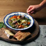 An image of a beautifully presented bowl of Welsh cawl on a wooden board with cheese and bread alongside