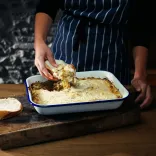 A roasting tin with freshly baked Anglesey eggs, with a chef's hand scooping some out on a slice of cut bread
