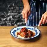 A beautifully presented bowl of faggots with mash, covered in gravy, being cut open for serving by a chef's hands