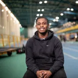 Jeremiah Azu sitting on an indoor athletics track, smiling at the camera.
